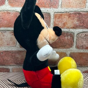 May include: A black and red plush Mickey Mouse toy sitting with its legs crossed. The toy has yellow shoes and a black tail.