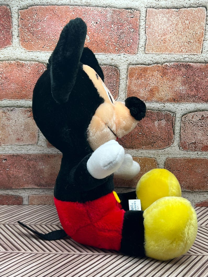 May include: A black and red plush Mickey Mouse toy sitting with its legs crossed. The toy has yellow shoes and a black tail.