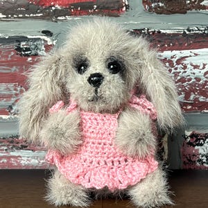 May include: A small, gray, furry toy dog wearing a pink crocheted dress. The dog has black eyes and a black nose.