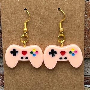 Puede incluir: Un par de pendientes de color rosa con forma de mando de videojuegos con ganchos dorados. Los mandos tienen botones negros, un corazón rojo y botones de colores.