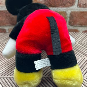 May include: Black and red plush Mickey Mouse toy with yellow shoes. The toy has a black tail and a tag that reads "All new materials - Polyester fibres.  Made in China.  Dist. by:  1218 W.  Mavis T.  CA.  Canada Trading Corp.  Burbank, CA.  Product # K134.  MTH-102."