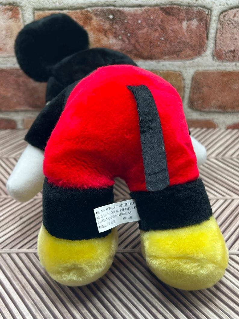 May include: Black and red plush Mickey Mouse toy with yellow shoes. The toy has a black tail and a tag that reads "All new materials - Polyester fibres.  Made in China.  Dist. by:  1218 W.  Mavis T.  CA.  Canada Trading Corp.  Burbank, CA.  Product # K134.  MTH-102."