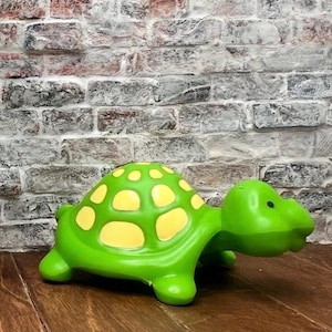 May include: A bright green turtle figurine featuring a smooth, rounded shell decorated with yellow oval spots. The turtle has a simple, molded head with a small black dot for an eye, four short legs, and a curved tail, all rendered in a solid, glossy finish.