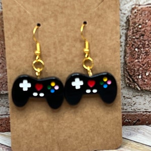 Puede incluir: Un par de pendientes negros y dorados con forma de mandos de videojuegos. Los mandos tienen botones blancos, un corazón rojo y botones de colores.