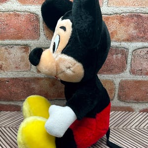 May include: A plush Mickey Mouse toy sitting with his legs crossed. He is wearing red shorts with black trim and yellow shoes. He has black ears and a black nose.