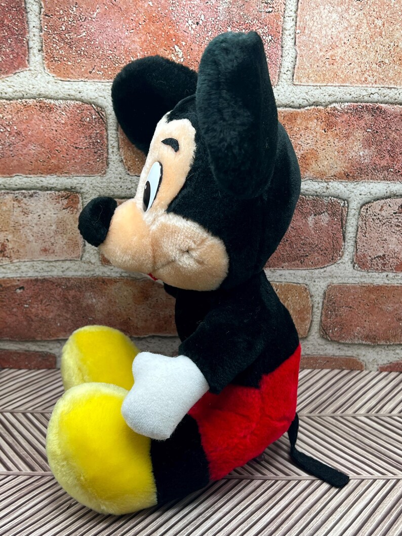 May include: A plush Mickey Mouse toy sitting with his legs crossed. He is wearing red shorts with black trim and yellow shoes. He has black ears and a black nose.