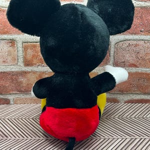 May include: A black and red plush Mickey Mouse toy sitting on a patterned surface. The toy has a yellow face and black ears.