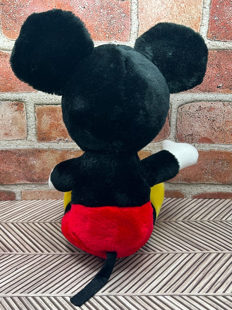 May include: A black and red plush Mickey Mouse toy sitting on a patterned surface. The toy has a yellow face and black ears.