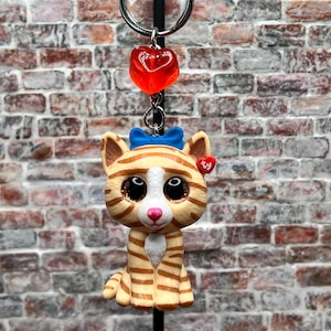May include: A keychain featuring a cartoon cat figurine with orange and tan stripes, a blue bow, and a red heart tag. The keychain also includes a red heart-shaped charm and a silver keyring.