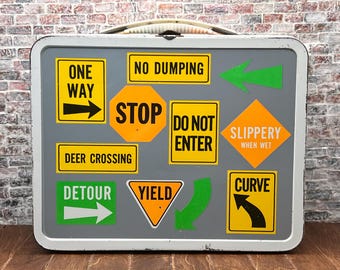 Puede incluir: Una fiambrera metálica vintage con una superficie gris, adornada con varias pegatinas de señales de tráfico. Las pegatinas incluyen "ONE WAY", "STOP", "DEER CROSSING" y "SLIPPERY WHEN WET". La fiambrera tiene un asa blanca y un borde plateado.