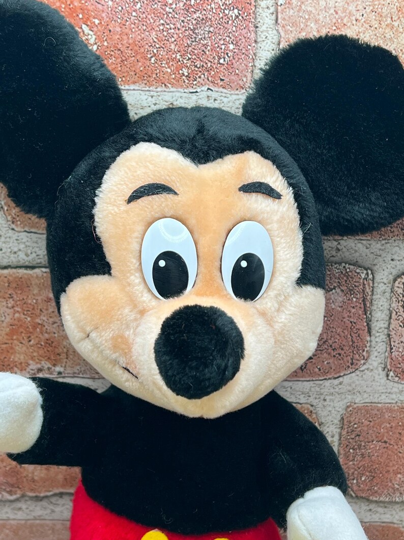May include: A plush Mickey Mouse toy with black ears, a black nose, and large white eyes. The toy is wearing red shorts with a yellow button.