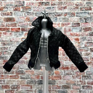 Könnte beinhalten: Eine schwarze Jacke mit Muster und Reißverschluss wird auf einer Schaufensterpuppe präsentiert. Die Jacke hat ein strukturiertes Design und einen Kragen. Die Schaufensterpuppe ist silberfarben und steht vor einer Backsteinmauer.