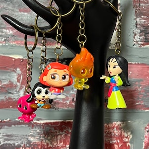 May include: Four colorful character keychains with gold chains. The characters include a pink octopus, a brown and white dog, a red-haired girl with glasses, and a yellow and orange fire creature. The keychains are hanging from a black display stand.
