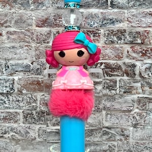 May include: A blue and pink pen with a doll-like topper. The doll has pink hair, a pink dress, and a blue bow. The pen has a fuzzy pink pom-pom at the bottom.