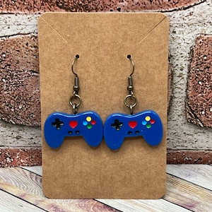 Puede incluir: Un par de pendientes azules de mando de videojuego con un corazón rojo y botones de colores. Los pendientes están en una tarjeta marrón.