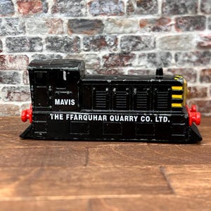 Op de afbeelding: Een zwart speelgoedtreinmodel met de naam "MAVIS". De trein heeft rode accenten en de tekst "THE FFARQUHAR QUARRY CO. LTD." is op de zijkant gedrukt. Geplaatst tegen een bakstenen muur.