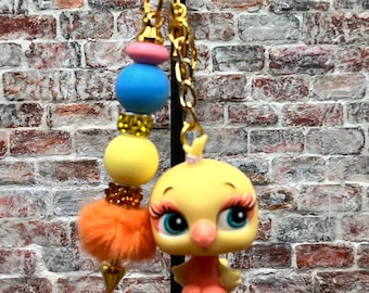 May include: A gold-colored keychain featuring a yellow chick figurine with large blue eyes and pink eyelashes. The keychain also includes a beaded charm with pink, blue, and yellow beads, an orange pom-pom, and a gold-colored spike.