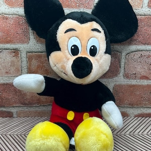 May include: A plush Mickey Mouse toy sitting with his arms outstretched. He is wearing his signature red shorts with yellow buttons and has large black ears and yellow shoes.