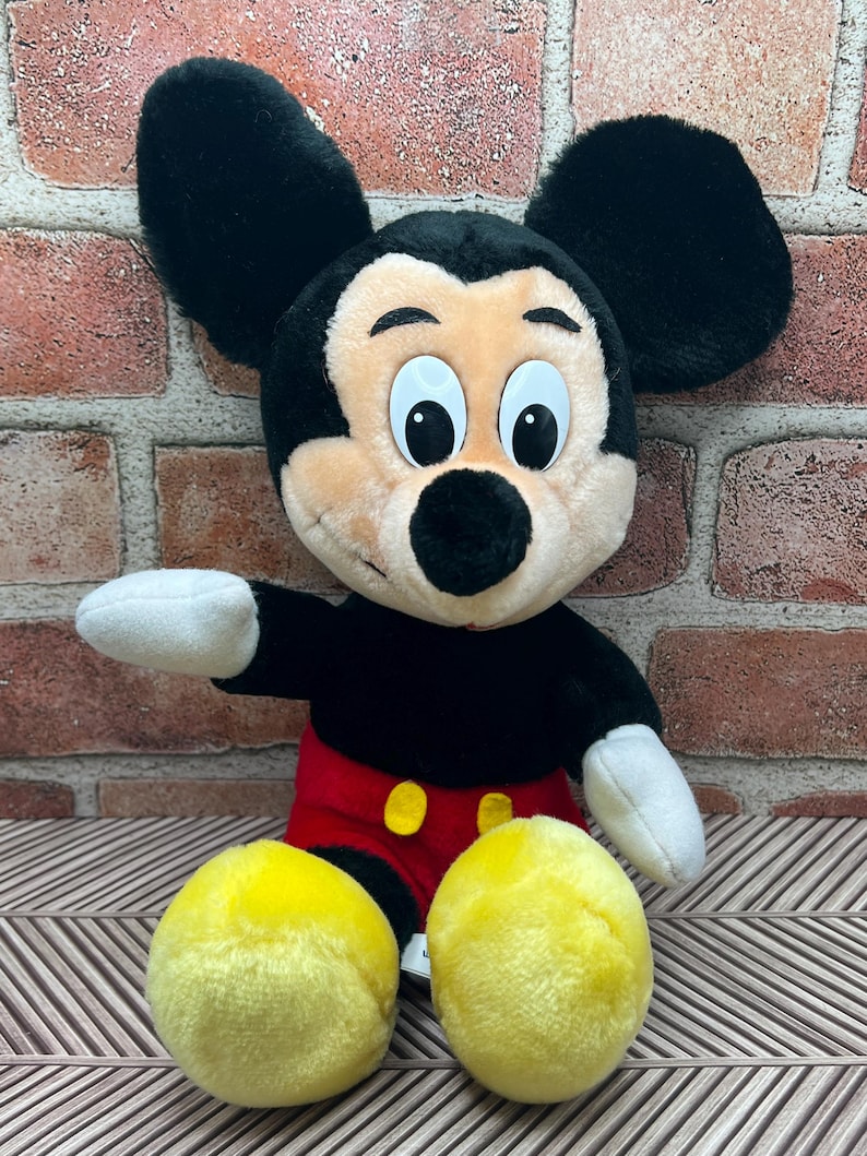 May include: A plush Mickey Mouse toy sitting with his arms outstretched. He is wearing his signature red shorts with yellow buttons and has large black ears and yellow shoes.