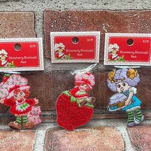 May include: Three embroidered iron-on patches featuring Strawberry Shortcake characters. The patches are on a brick background. The first patch shows Strawberry Shortcake holding a cat. The second patch shows Strawberry Shortcake holding a strawberry. The third patch shows a purple-haired character holding a cake.