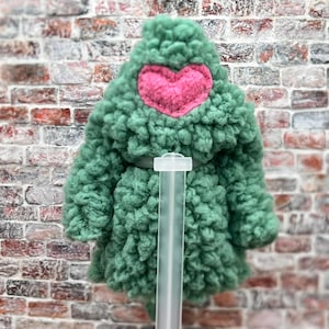 Op de afbeelding: Een groen, getextureerd fleecejack met capuchon, voorzien van een roze hartvormige patch in het midden van de capuchon. Het jack wordt gepresenteerd op een doorzichtige plastic standaard, wat het dikke, krullende materiaal benadrukt.