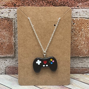 May include: A silver chain necklace with a black plastic pendant shaped like a video game controller. The controller has white and red buttons, and colorful buttons on the right side.