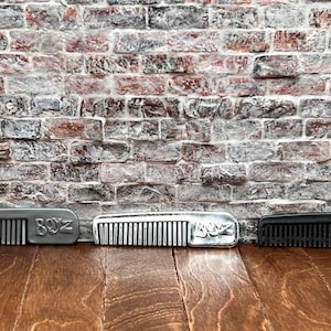 May include: Three combs in a row, each with the text "BOYZ". The combs are in silver, silver, and black. The combs are on a wooden surface in front of a brick wall.