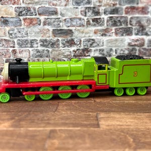 Op de afbeelding: Een speelgoedtrein met een groene locomotief, een zwarte schoorsteen en een rood chassis. De trein heeft groene wielen en een kolenwagen. Het nummer 3 is zichtbaar op de zijkant van de kolenwagen. Het speelgoed staat tegen een bakstenen muur.