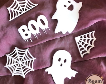 Może przedstawiać: Pięć białych dekoracji halloweenowych na różowym tle z tkaniny. Dekoracje obejmują dwa duchy, dwie pajęczyny i słowo "BOO" z kapiącą farbą.