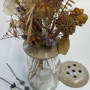 May include: A clear glass vase with a beige ceramic lid holds a dried flower arrangement. The arrangement includes beige, brown, and purple dried flowers. A matching ceramic flower frog sits beside the vase. Lavender sprigs are in the foreground.