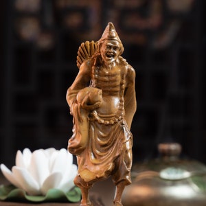 May include: A wooden figurine of a smiling figure wearing a hat and holding a round object. The figurine is carved with intricate details and has a warm brown color.