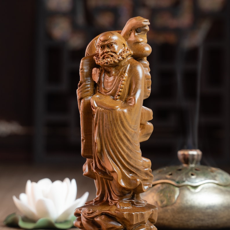 May include: A wooden sculpture of a Buddhist monk holding a staff and a scroll. The monk is wearing a robe and has a long beard. The sculpture is carved in detail and has a warm brown color.
