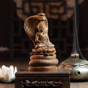 May include: A wooden sculpture of Buddha seated on a lotus flower, with a cobra hood behind him. The sculpture is brown and features a detailed design. It is placed on a wooden base.