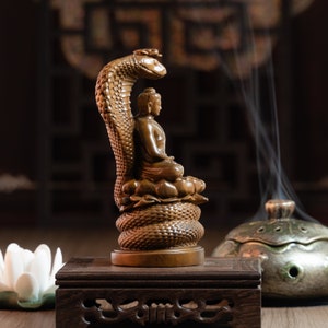 May include: A wooden statue of Buddha seated on a coiled snake, with a dark brown wooden base. The statue is on a wooden table with a small white flower in the foreground.