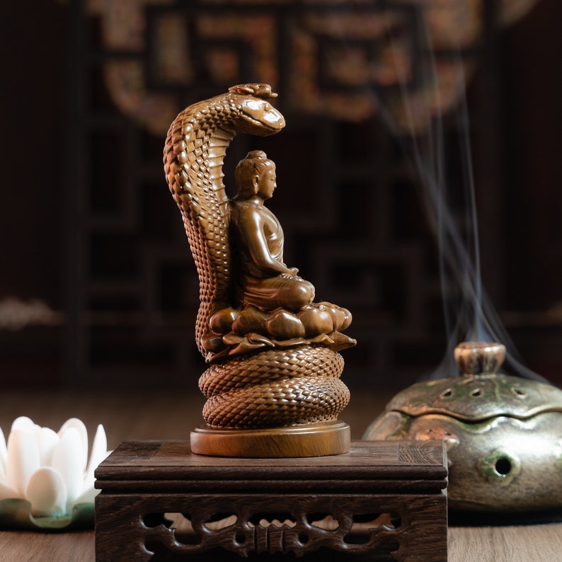May include: A wooden statue of Buddha seated on a coiled snake, with a dark brown wooden base. The statue is on a wooden table with a small white flower in the foreground.