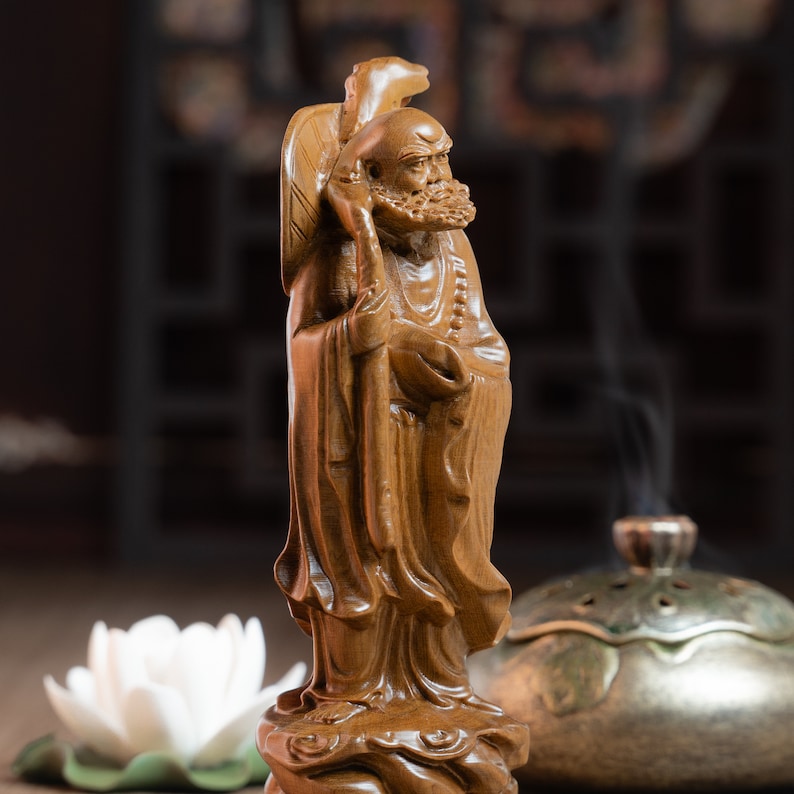 May include: A wooden sculpture of a Chinese deity, with a long beard and a staff, standing on a base with intricate carvings. The sculpture is brown and has a natural wood finish.
