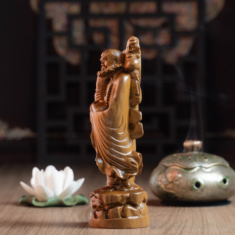 May include: A wooden statue of a Buddhist monk standing on a base with a tree behind him. The statue is carved in detail and has a warm brown color.