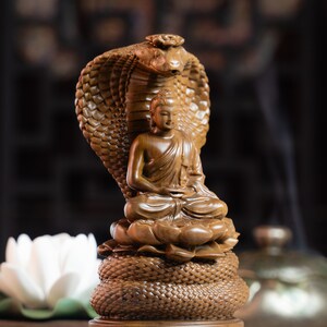 May include: A wooden sculpture of Buddha seated in meditation, with a cobra hood behind him. The sculpture is carved with intricate details and is a beautiful example of traditional Buddhist art.