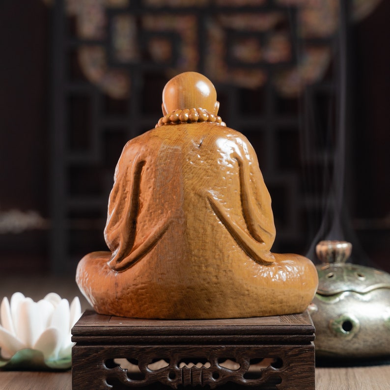 May include: A wooden figurine of a seated Buddha, carved in a detailed style, with a smooth, polished finish. The figurine is placed on a wooden stand with intricate carvings.