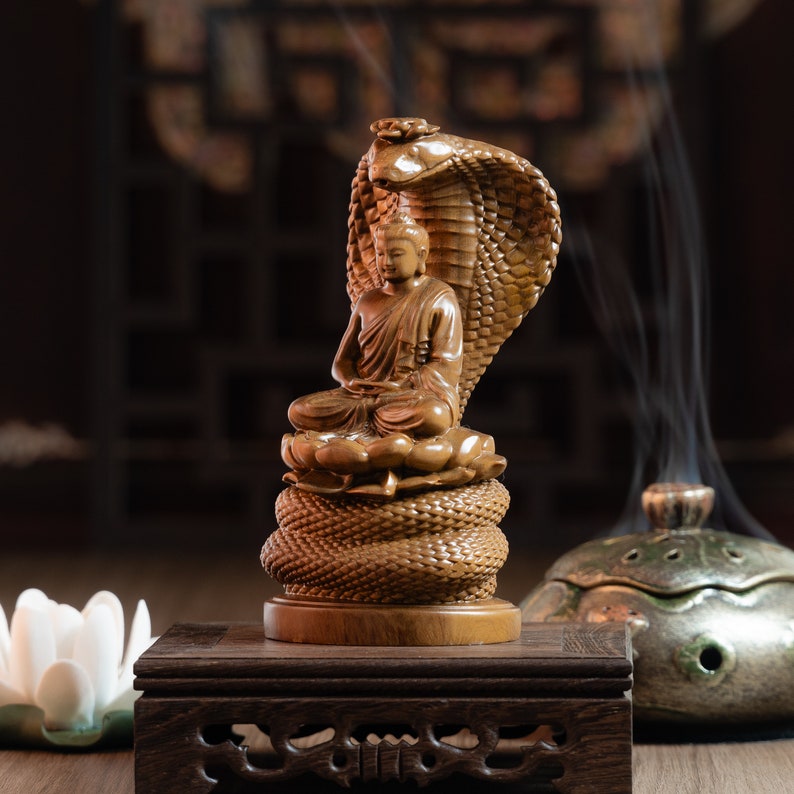May include: A wooden sculpture of Buddha seated on a coiled snake, with a lotus flower base. The sculpture is on a wooden platform with intricate carvings.