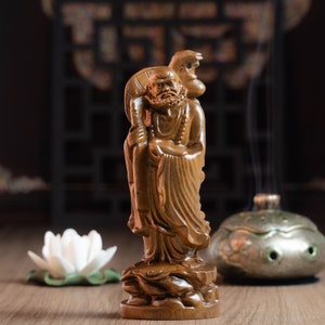 May include: A wooden statue of a man with a long beard and a staff, holding a small animal on his shoulder. The statue is standing on a base with a carved design. The statue is brown and has a natural wood finish.