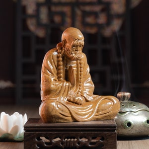 May include: A wooden statue of a meditating figure with a beaded necklace, sitting on a wooden base. The statue is brown and has a detailed face.