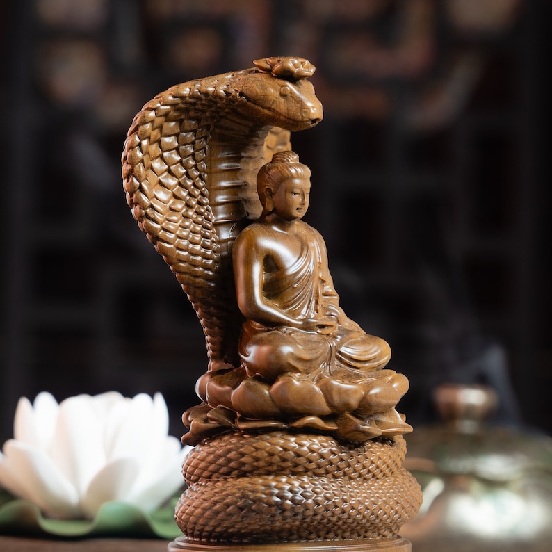 May include: A wooden sculpture of Buddha seated on a coiled snake, with the snake's head forming a canopy over Buddha's head. The sculpture is carved in a detailed, realistic style, with intricate details on the snake's scales and Buddha's robes.