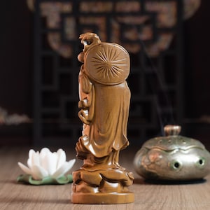 May include: A wooden statue of a standing figure with a circular halo behind their head. The statue is carved with intricate details and has a warm brown color.