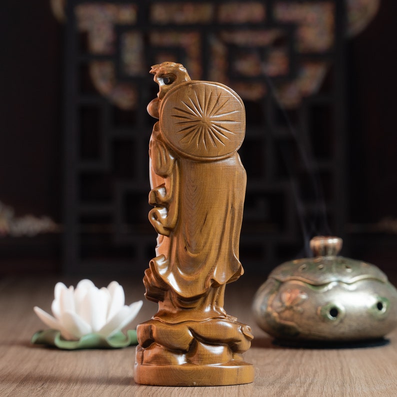 May include: A wooden statue of a standing figure with a circular halo behind their head. The statue is carved with intricate details and has a warm brown color.