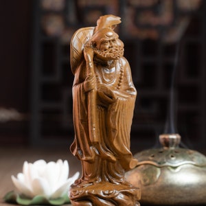 May include: A wooden statue of a smiling, bearded man holding a staff. The statue is carved in a traditional style and is wearing a long robe. The statue is standing on a base with a decorative design.
