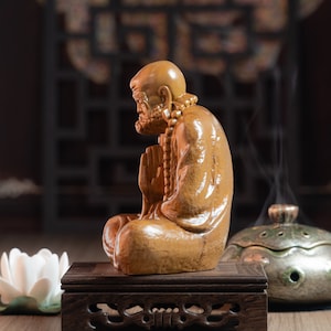 May include: A wooden statue of a meditating figure with a beaded necklace, sitting on a dark wood stand. The statue is carved in a realistic style and has a warm brown color.
