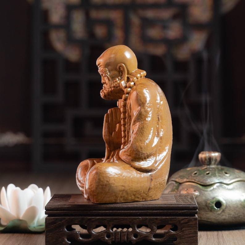May include: A wooden statue of a meditating figure with a beaded necklace, sitting on a dark wood stand. The statue is carved in a realistic style and has a warm brown color.