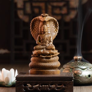 May include: A wooden statue of Buddha seated in meditation, with a cobra hood behind him. The statue is on a wooden base and is surrounded by incense smoke.