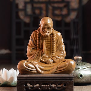 May include: A wooden statue of a meditating figure with a long beard and a beaded necklace. The statue is sitting on a wooden base with intricate carvings.